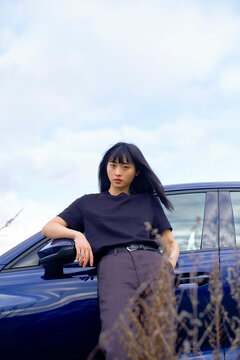 Woman in black shirt leans on a dark blue car against a cloudy sky backdrop. Brandenburg, Germany
