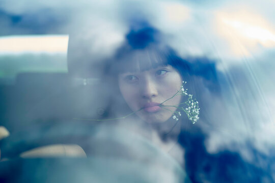 Woman with flower in mouth gazing through a car window with cloud reflections. Brandenburg, Germany