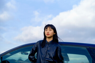 Young woman in a leather jacket stands by a car under a clear blue sky. Brandenburg, Germany