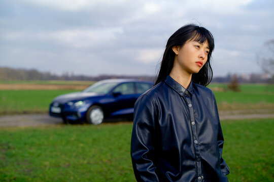 Woman in a leather jacket stands in front of a parked car in a vast green field. Brandenburg, Germany
