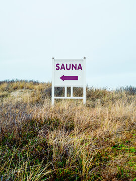 A sign with a purple arrow points to a sauna amidst dry grass and sandy dunes. Sylt, Germany