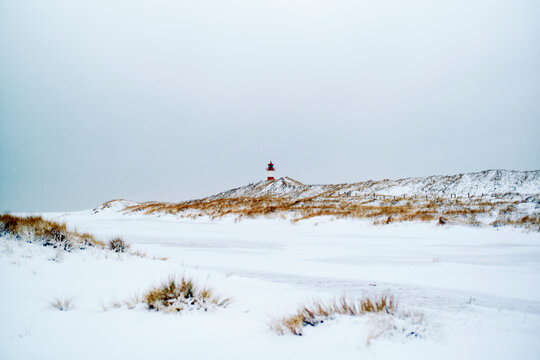 Snow-covered landscape with distant lighthouse on a hill under overcast sky. Sylt, Germany