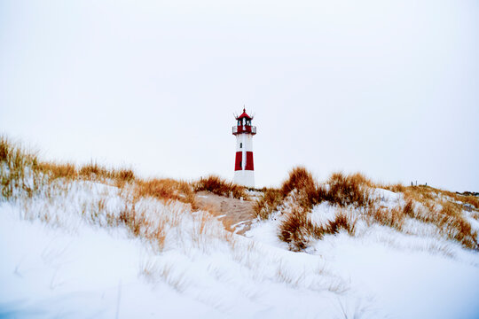 A red and white lighthouse stands amid snow-covered dunes under a bright sky. Sylt, Germany