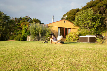 Woman relaxing on a wooden chair in a sunny garden with a house and lush greenery. Waiheke Island, Auckland, New Zealand