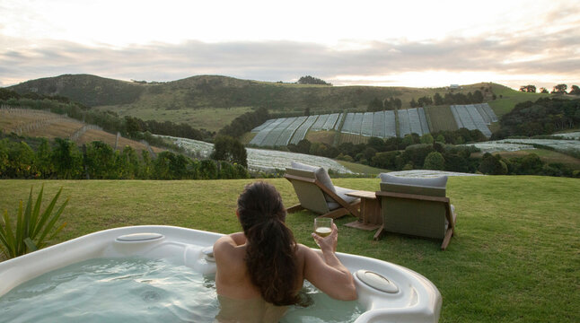 Woman relaxing in a hot tub with wine, overlooking scenic vineyard landscape at sunset. Waiheke Island, Auckland, New Zealand