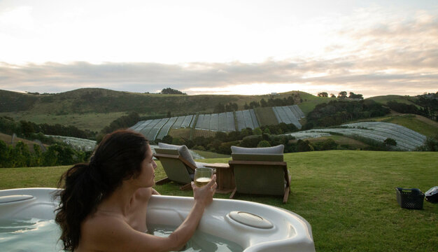 Woman in hot tub holding a wine glass, overlooking vineyards on a hill during sunset. Waiheke Island, Auckland, New Zealand