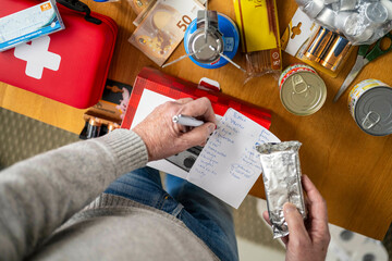 Person makes list of emergency supplies on wooden table with canned goods and red first aid kit. Preparing a survival weekend