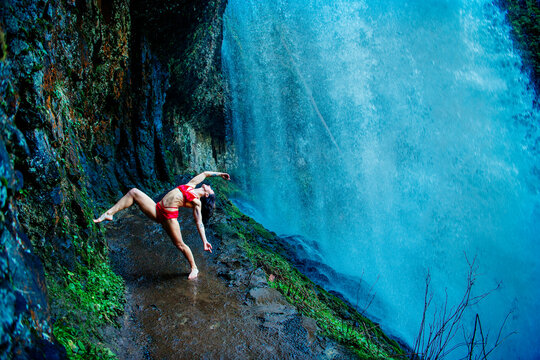 Dancer in red poses gracefully by a stunning waterfall on a rocky and wet path. Silver Falls, Oregon, USA