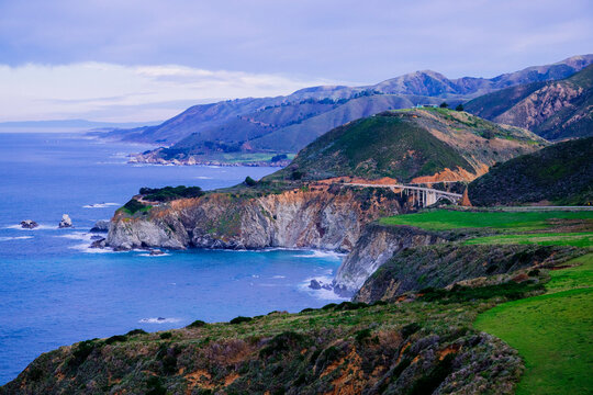 A stunning coastal landscape with rugged cliffs, the famous Bixby Creek Bridge, and vibrant blue ocean waves. California, USA