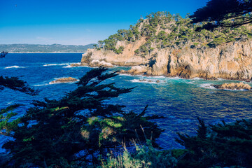 Rocky coastline with blue sea, distant hills, and lush trees under a clear sky. Point Lobos State Natural Reserve, CA, USA