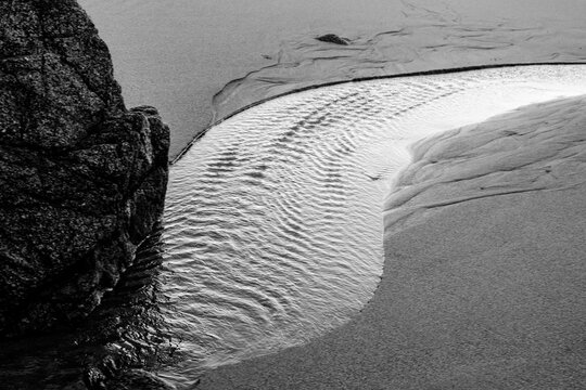Black and white image of textured sand and water near a large rock on a beach. Monterey, CA, USA