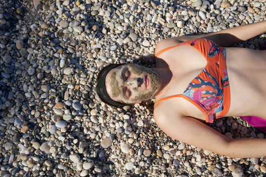 Woman in a bikini with a mud face mask, relaxing on a pebbly beach.