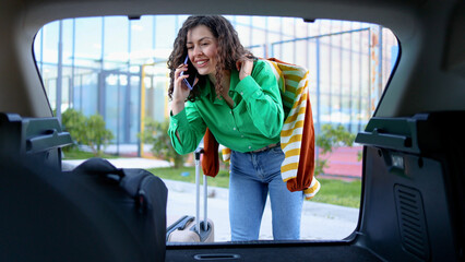 Young woman loading luggage in car trunk, smartphone pressed to ear, readying for travel