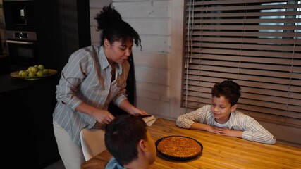 In a kitchen, a mixed-race mother in her 30s talks while cutting a pie with a knife on a wooden table, as her two school-aged sons sit and watch her, all casually dressed, next to a counter and blinds
