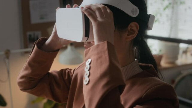 Asian female developer putting on VR headset, interacting in 3D environment while testing virtual reality application in office