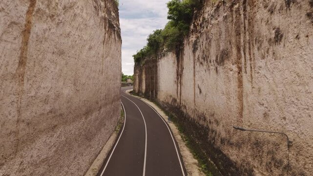 Cinematic low aerial shot gliding above a narrow, empty road between towering stone cliffs at Tanah Barak, Bali, Indonesia&mdash;showcasing stunning natural formations and peaceful tropical scenery.