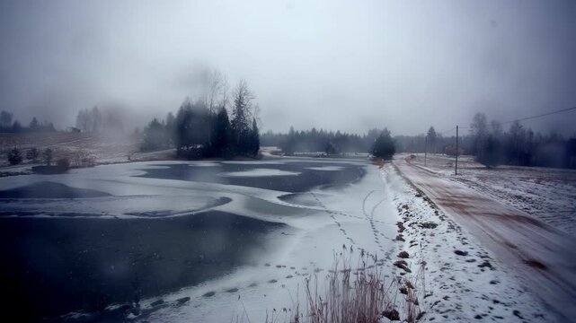 Heavy snow descends over a partially frozen lake and rural road near Indrani Guesthouse in Skujene, Latvia, captured during a timelapse.