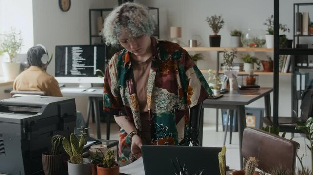 Young female employee with prosthetic arm taking document from printer, then walking to desk, examining it and using laptop during workday in modern office