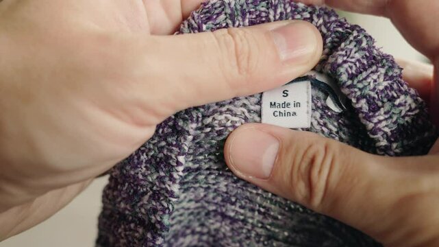 Close-up of hands holding a black clothing label marked "Made in China" inside a gray garment, reflecting global textile manufacturing and political tensions over tariffs and trade restrictions.