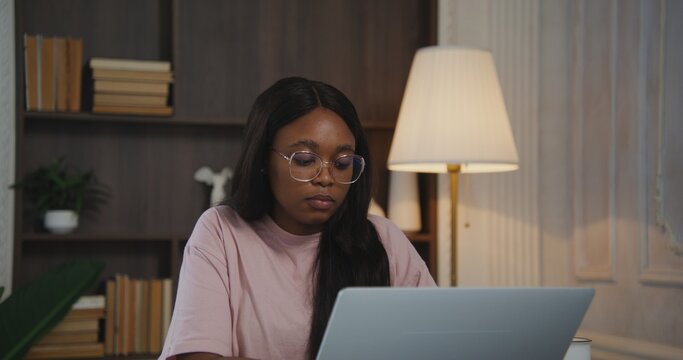 Beautiful young african american woman in glasses works on a laptop making notes in a notepad sitting at a desk in a modern stylish interior