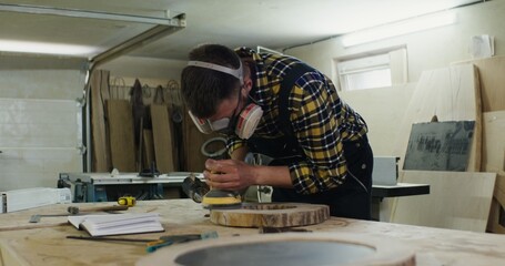 a man of European appearance in protective headphones and a mask processes a wooden saw cut using a tool, sanding wood with a flex, work in a carpentry workshop
