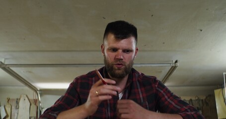 a serious man of European appearance with a beard, in a plaid shirt, is actively talking while looking at camera and gesturing with his hands in a carpentry workshop, a lot of wooden products