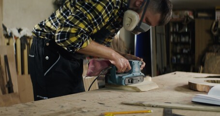 a man of European appearance in protective headphones and a mask processes a wood using a tool, sanding wood with a flex, work in a carpentry workshop