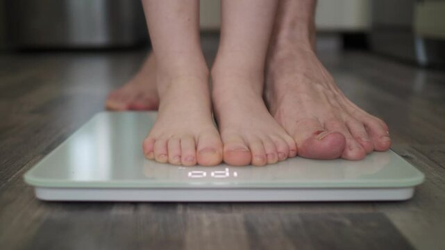 Children's and Men's feet step on scales to determine weight together with kid. Father and son look at total weight. Approaching from afar, out of focus. Home environment. Calm daytime shades of gray.