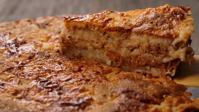 A single slice of golden brown apple puff pastry pie is carefully lifted upwards from the rest of the pie, revealing its flaky layers and the baked apple filling. 