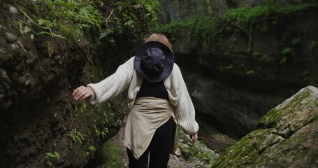 Russia, Caucasus. A young beautiful woman of European appearance, dressed in a warm sweater, makes her way through the stones in the cleft between the rocks overgrown with greenery