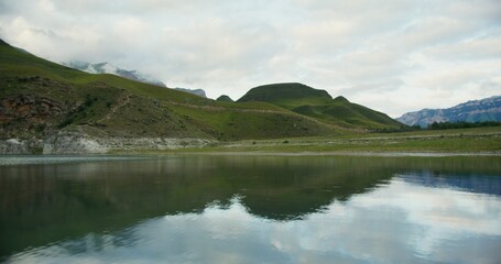 A beautiful view of the mountain lake at the foot of the densely vegetated hills. The tops of the rocky mountains are covered with thick clouds.