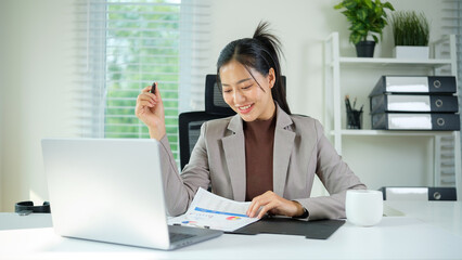 Happy young professional woman looks at printed charts while holding a pen at her bright, modern desk.