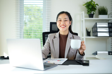 A confident and approachable businesswoman takes a moment to smile while sipping coffee in a light-filled office.