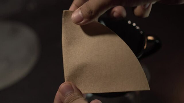 Folding a paper filter for a V60 coffee dripper on a black table with black brewing equipment. Minimalist and moody barista preparation scene.