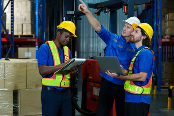 Three warehouse workers wearing safety helmets and vests discussing logistics operation using tablet and laptop inside industrial storage with teamwork and cooperation during duty
