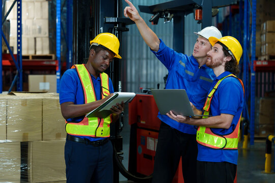 Three warehouse workers wearing safety helmets and vests discussing logistics operation using tablet and laptop inside industrial storage with teamwork and cooperation during duty