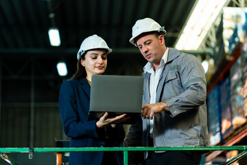 Supervisor and secretary reviewing warehouse data on laptop while collaborating on logistics task inside warehouse environment with professional attire and safety helmets