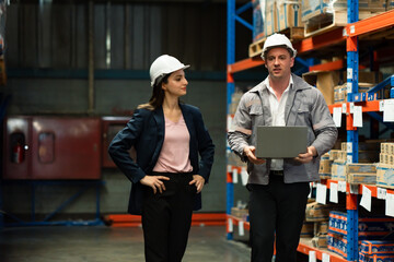 Caucasian male warehouse manager walking with female secretary during warehouse inspection holding laptop and having discussion while supervising shelf stock in industrial aisle