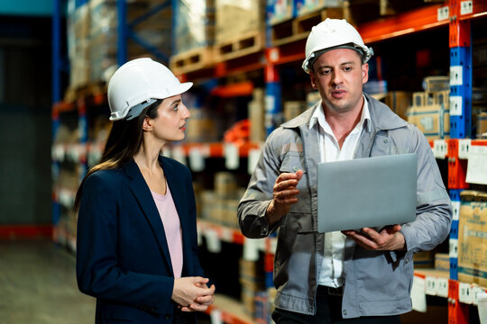 Warehouse manager holding laptop and explaining work process to secretary while discussing supply chain task inside stockroom during team planning with safety helmets and formal wear