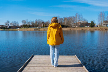 Fototapeta premium Woman in yellow jacket and jeans standing on wooden dock by lake, thinking quietly, countryside houses across the water, sunny spring day, peaceful mood, solitude, calm nature