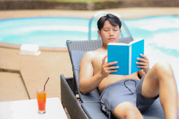 Asian man relaxing by the pool, enjoying a captivating book and sipping a refreshing cocktail under the bright sun on a perfect vacation day. Embracing leisure and tranquility