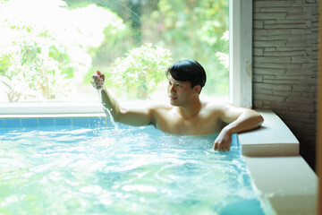 Asian man relaxing in a hot tub, playfully splashing water while enjoying a serene view of lush greenery through the window in a tranquil spa setting