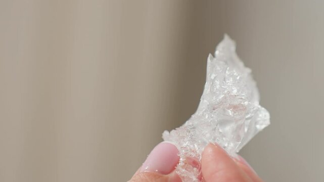 Close up of delicate hand with pink manicured nails gently shaking transparent rubber-like soluble material, highlighting intricate texture and clarity, set against soft neutral background