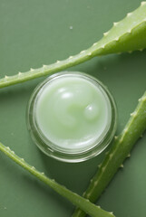 Top-down product photo of light green cream in glass jar with aloe vera leaves on a muted green background. Natural beauty, spa concept. Minimalist, clean style.