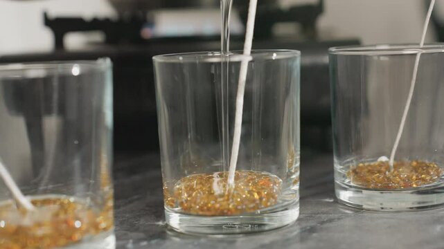 Close up of candle wax production process showing person pouring hot melted wax into clear glass cup filled with golden wax beads and wick centered inside