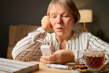A woman sits at a wooden table, intently reading a message on her phone. She appears surprised, with a cup of tea and some cookies nearby, creating a warm atmosphere