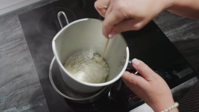 Female chandler melts tallow flakes inside metal mixing bowl placed in pot of hot water on electric stove, using double boiler method to safely liquefy wax substance for candle making in home kitchen