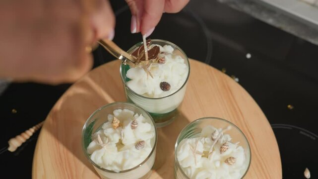 House wife trims long wick in center candle jar using golden wick trimmer while holding it steadily, surrounded by two more decorative wax jars on wooden tray adorned with miniature shells