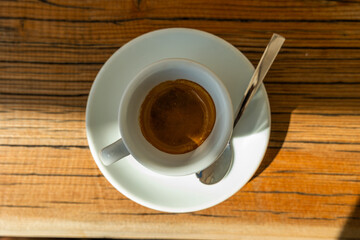 primo piano dall'alto su una tazzina con caffè espresso, su un piatto, con un cucchiaio piccolo, su un tavolo di legno, di giorno, a pranzo, illuminato a metà dal sole