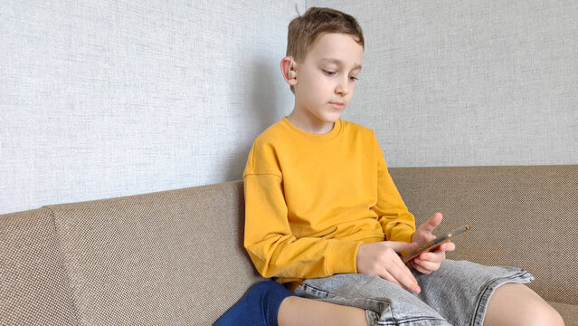 Boy with hearing aid in ear holds phone in hands, listens to audiobook while sitting in his room